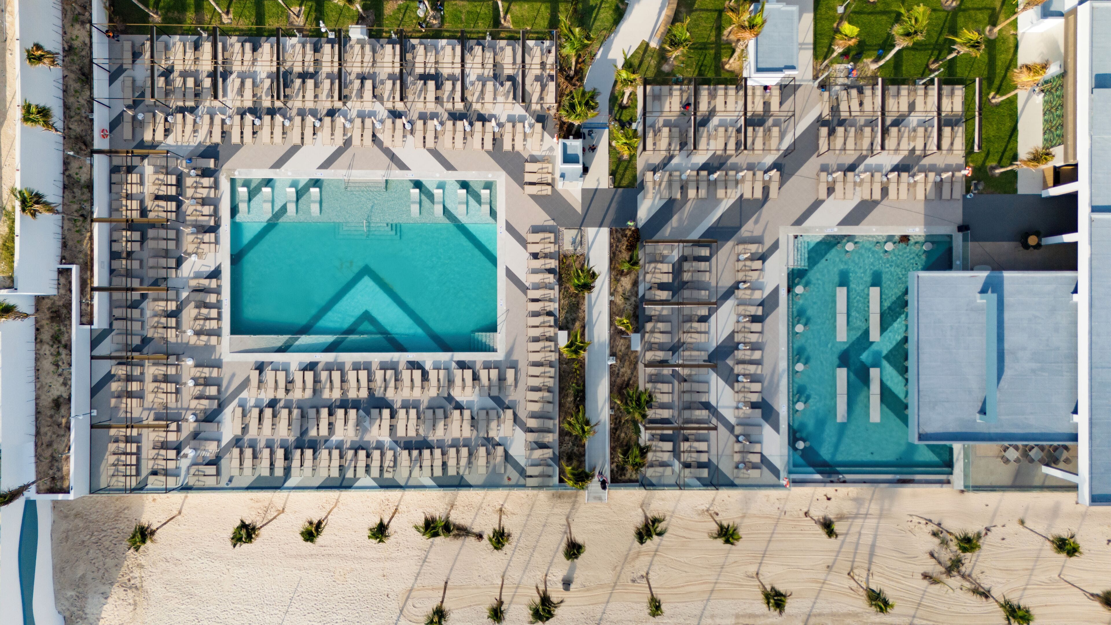 Vue aérienne d’un complexe hôtelier doté de deux grandes piscines, de chaises longues et d’une plage de sable.
