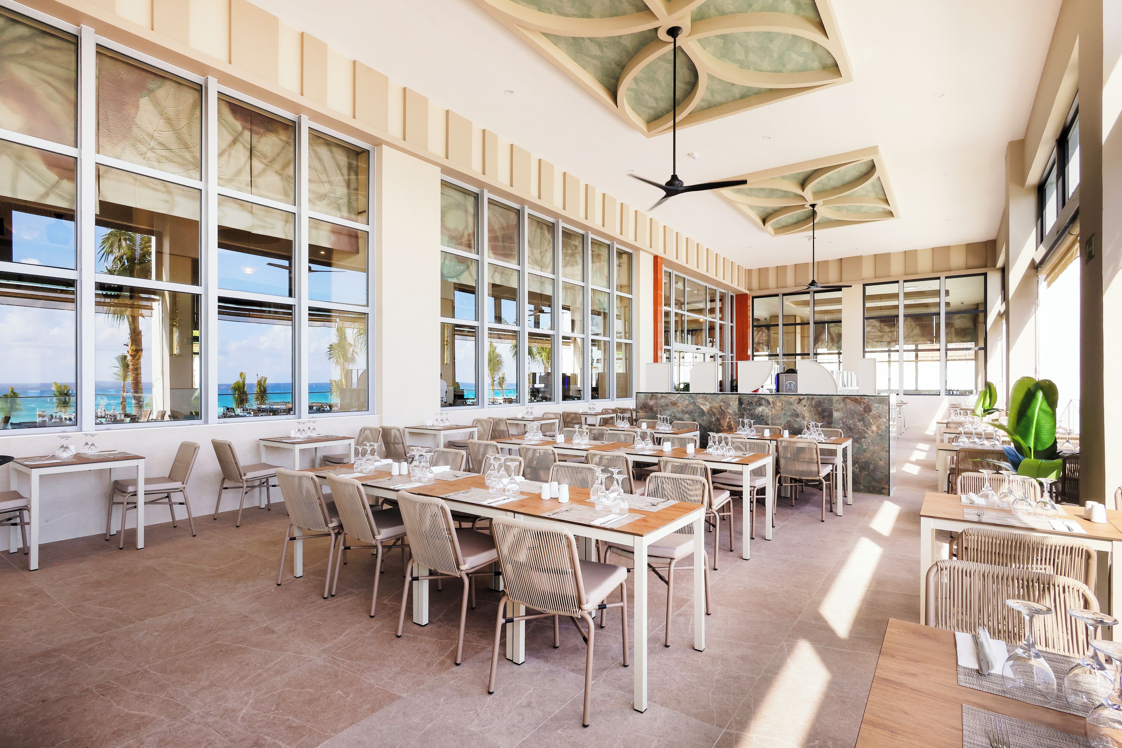 Un comedor amplio con varias mesas y sillas, ventanas grandes que ofrecen vistas al océano y un ventilador de techo.