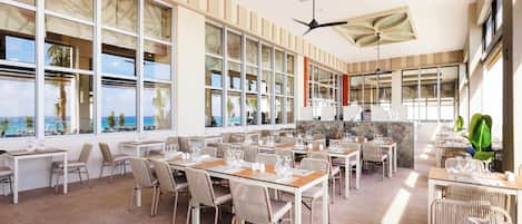 A spacious dining area with multiple tables and chairs, large windows offering views of the ocean, and a ceiling fan.