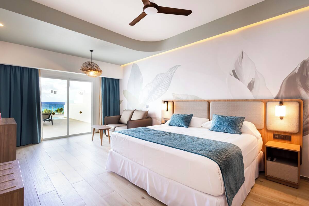 Una habitación de hotel moderna con una cama grande, un ventilador de techo, una mesita de noche y vistas al océano a través de la puerta corredera de vidrio.