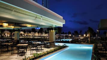 An outdoor pool area with tables and chairs, surrounded by palm trees and a pool.