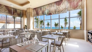 Un comedor amplio con mesas dispuestas para los huéspedes, ventanas grandes que ofrecen vistas a palmeras y al océano, y un diseño interior moderno.