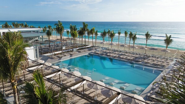 A resort pool with sun loungers and umbrellas, surrounded by palm trees and a view of the ocean.