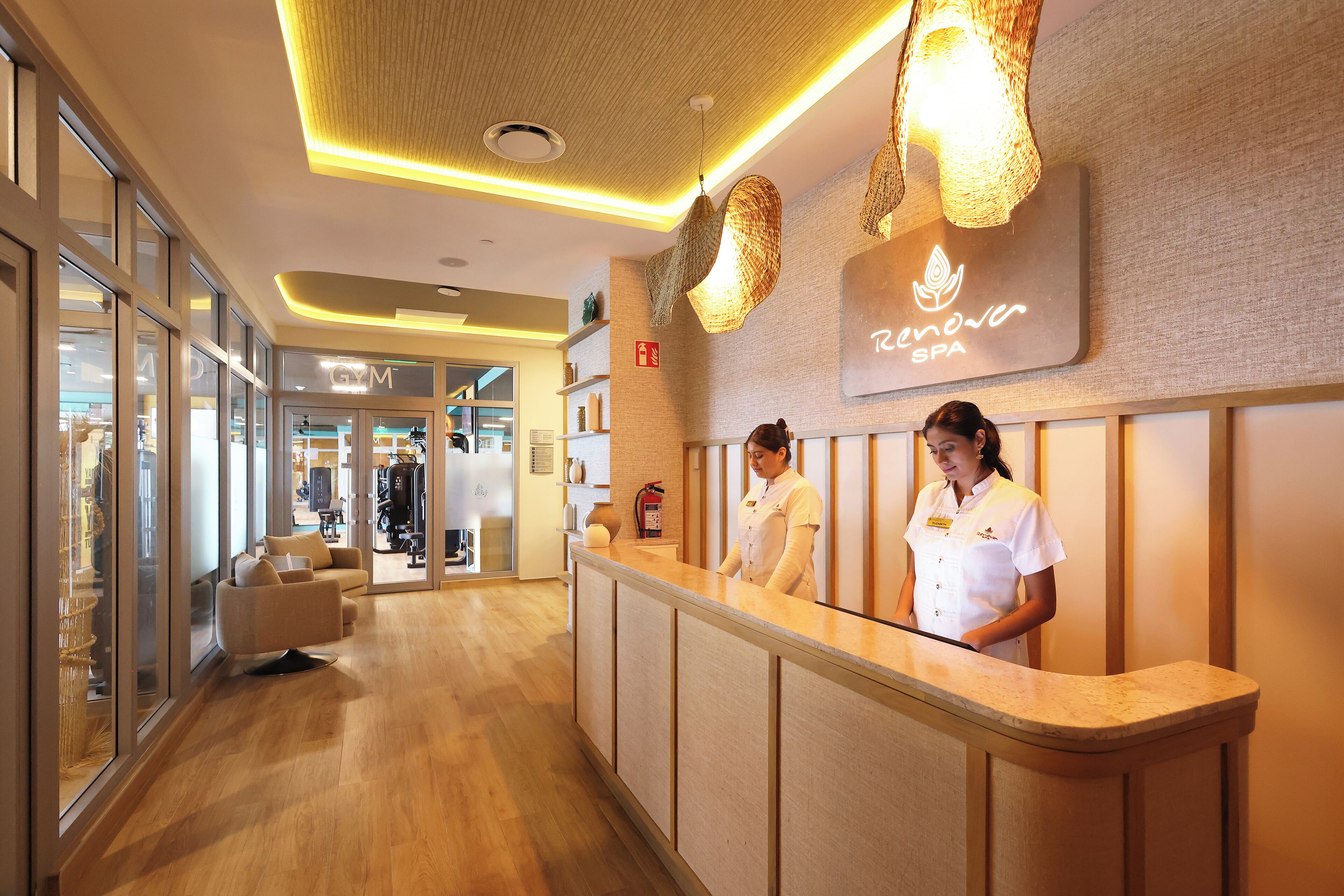 Un espace d’accueil avec un comptoir de réception, deux employés, une enseigne indiquant « Renovo Spa », et, au fond, on aperçoit la salle de sport.