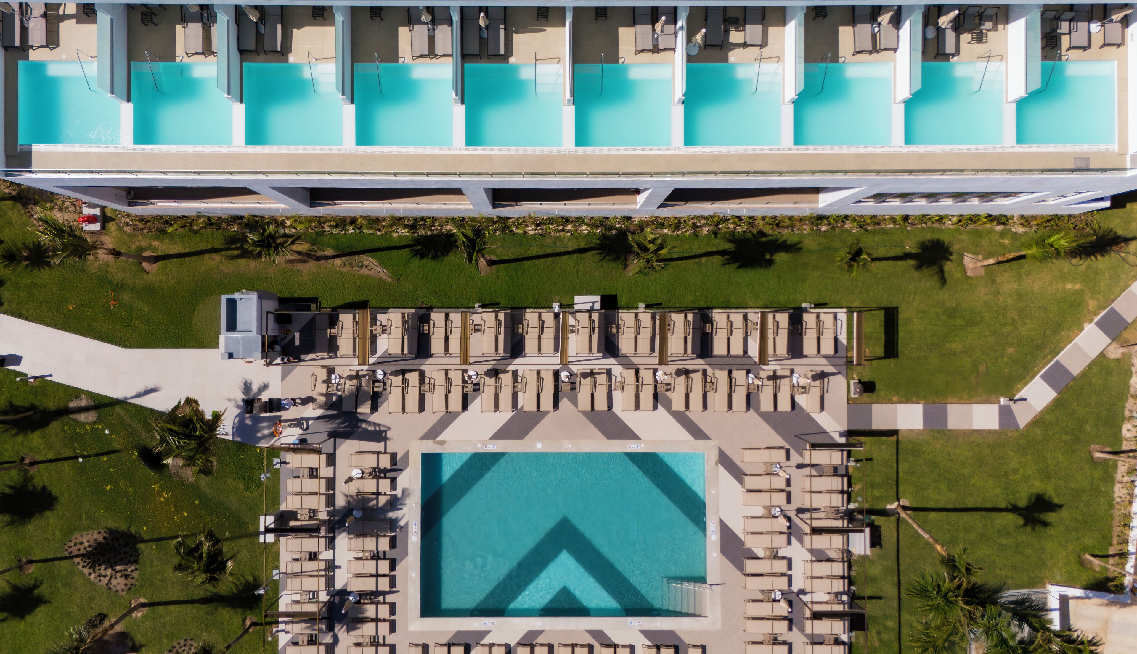 Vista aérea de un hotel con múltiples piscinas, tumbonas y una piscina central con un diseño geométrico distintivo.
