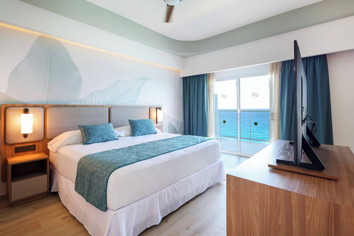 Habitación de hotel moderna con una cama grande, un escritorio y un televisor de pantalla plana, y un balcón con vistas al mar.