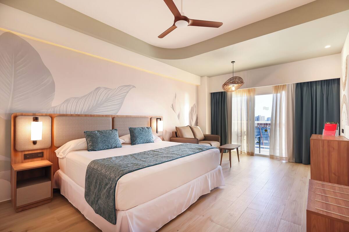 Habitación de hotel moderna con una cama grande, ventilador de techo, mesita de noche, escritorio y vistas de la ciudad desde el balcón.