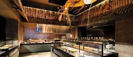 A buffet area with a variety of food items displayed in glass cases, a central island with seating, and a modern ceiling design with hanging elements.