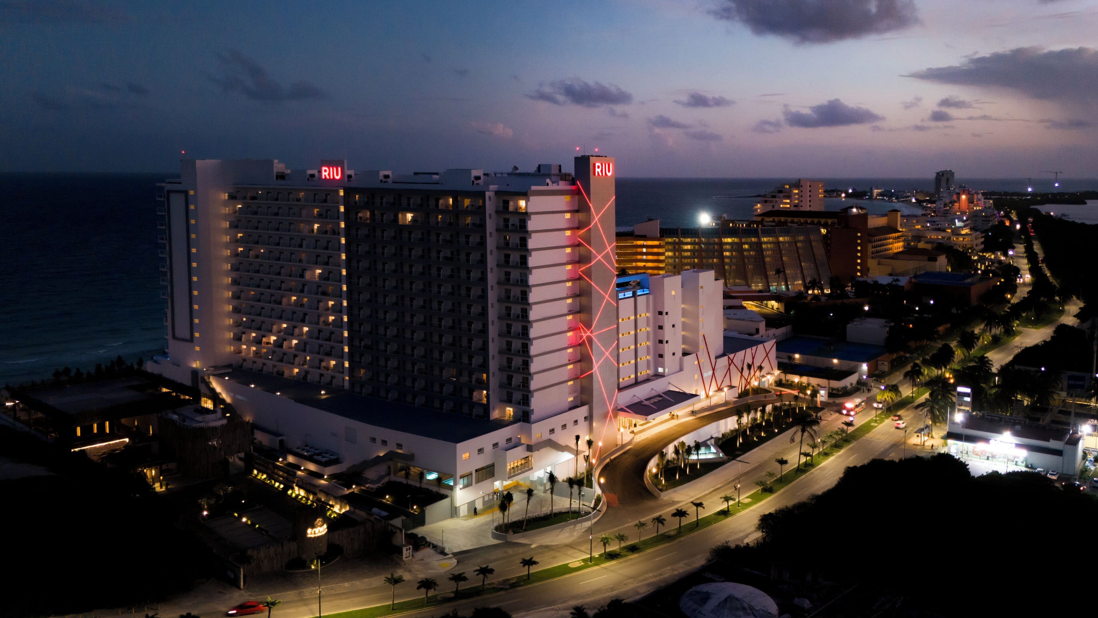 Un hôtel à plusieurs étages, reconnaissable à son grand panneau rouge « RIU », situé au bord d’une route côtière et entouré d’autres bâtiments.