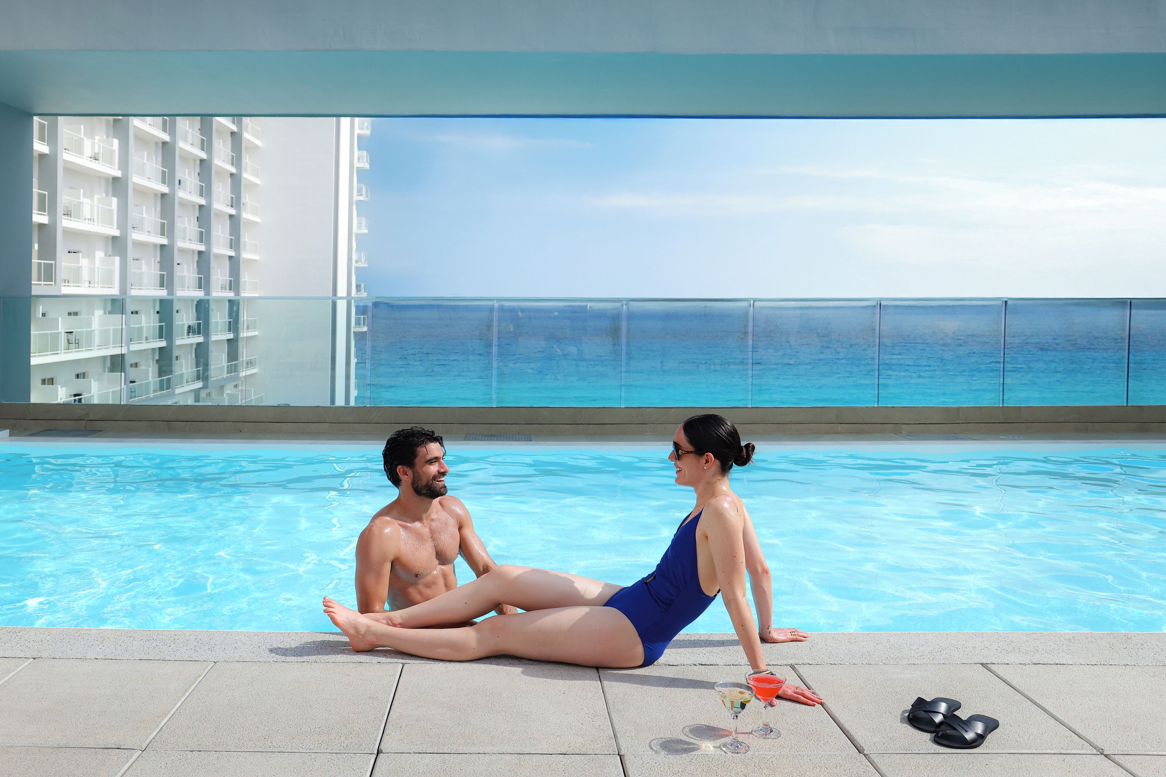 Una pareja junto a una piscina con vistas al océano y un edificio al fondo.