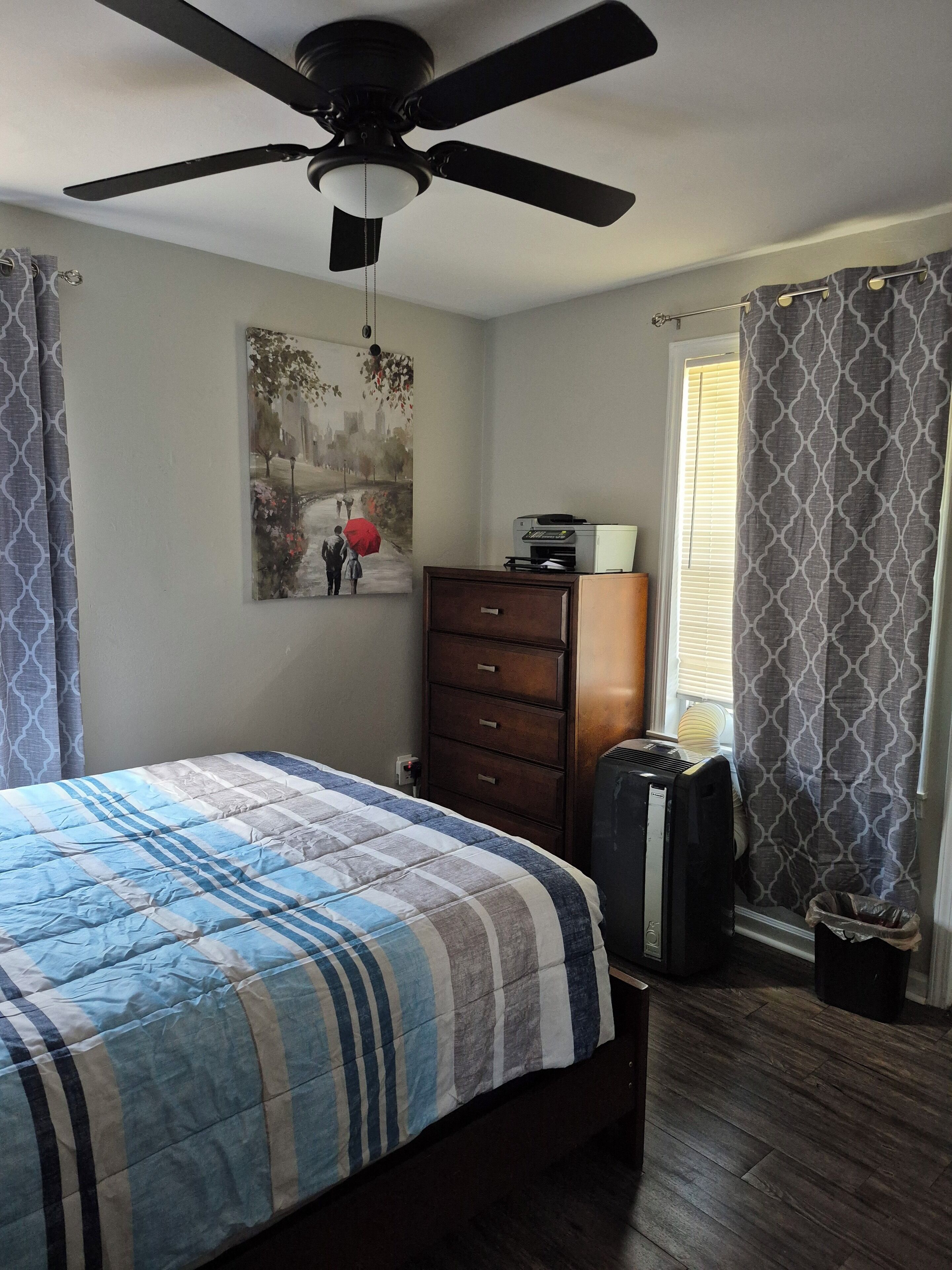 A bedroom with a bed, a ceiling fan, a dresser, a radiator, and a window with curtains.
