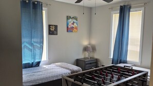 Un dormitorio con cama, ventilador de techo, ventana con cortinas, un cuadro en la pared y una mesa de futbolín.