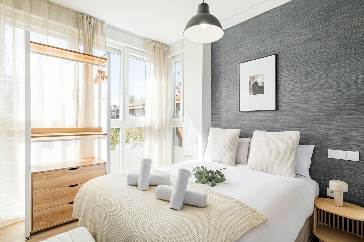 Un dormitorio moderno con una cama grande, mesitas de noche, una ventana con cortinas y una pared gris con textura.