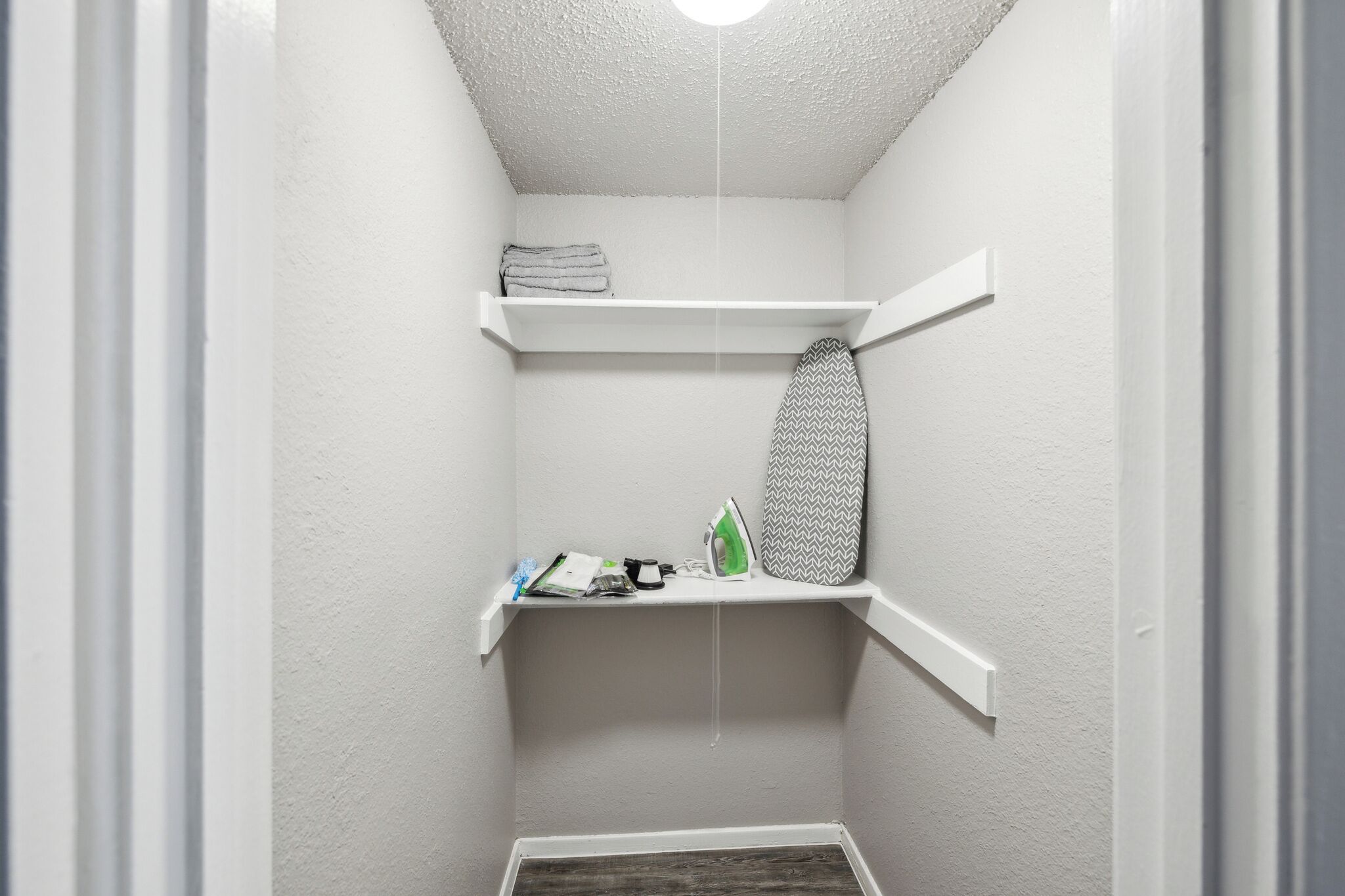 A small, narrow laundry room with white walls and a wooden floor. There are two white shelves, one above the other, with a folded towel on the top shelf and an iron on the bottom shelf.