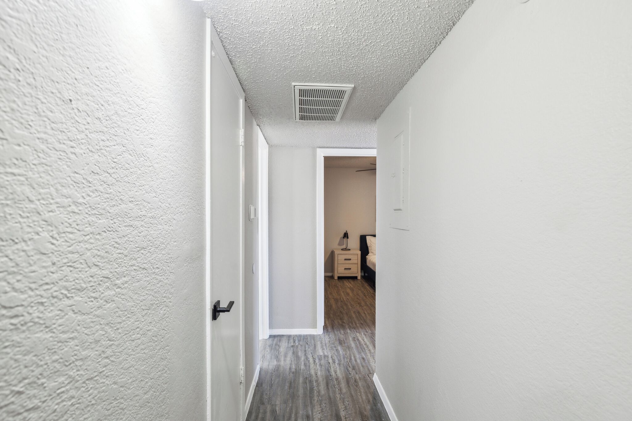 Een hal met een witte, geribbelde wand, een deur links en een bed met nachtkastje aan het einde van de kamer.