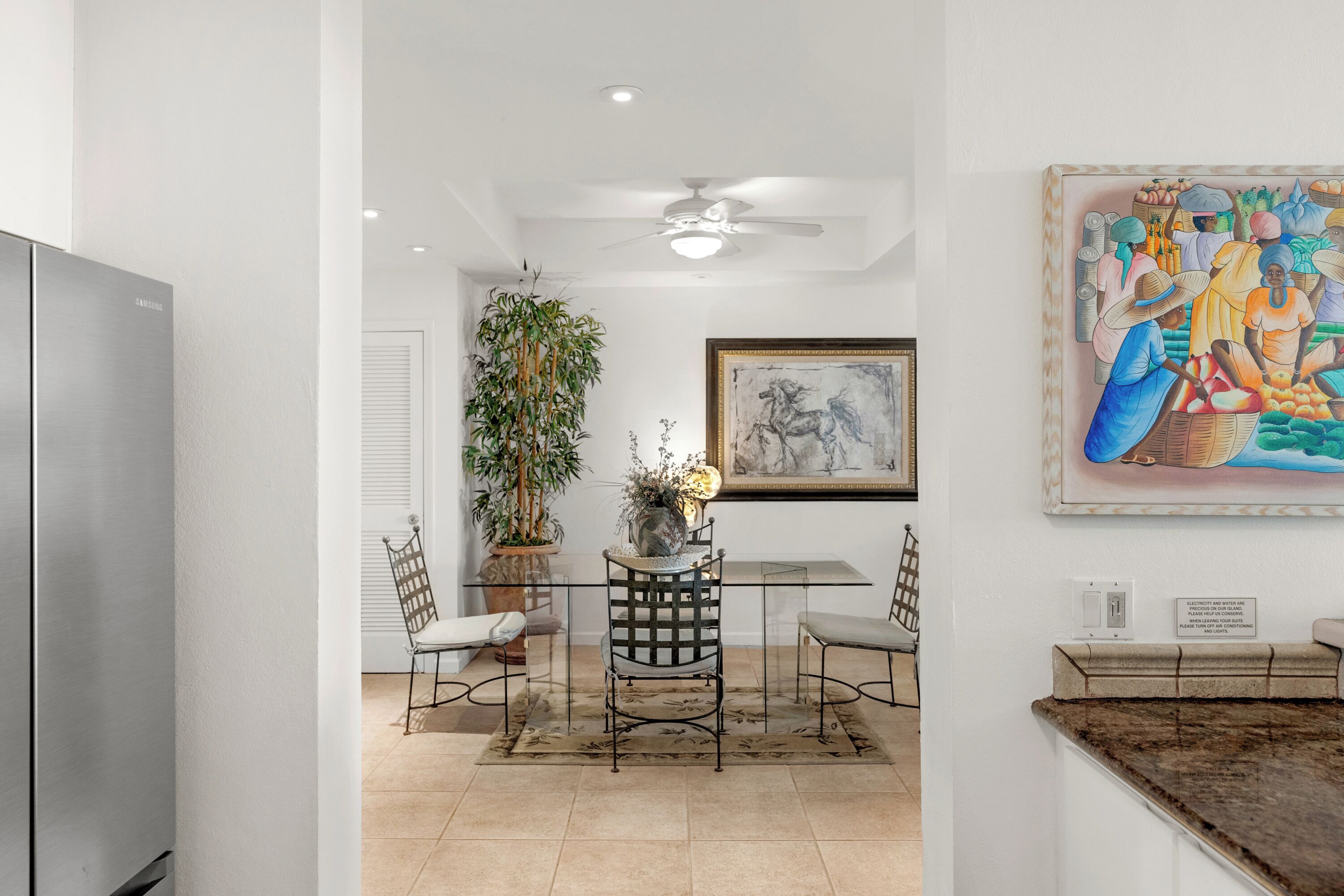 A modern kitchen with a stainless steel refrigerator, a dining area with a glass table and metal chairs, a ceiling fan, a large potted plant, and a framed painting on the wall.