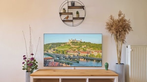 Un televisor sobre un soporte de madera, con un cuadro de paisaje urbano encima, un estante montado en la pared con objetos decorativos, y un florero con plantas secas.