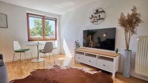 Un salón moderno con televisor de pantalla plana, una mesa de comedor con sillas, un tapete grande de cuero de vaca y una ventana con vista a follaje.