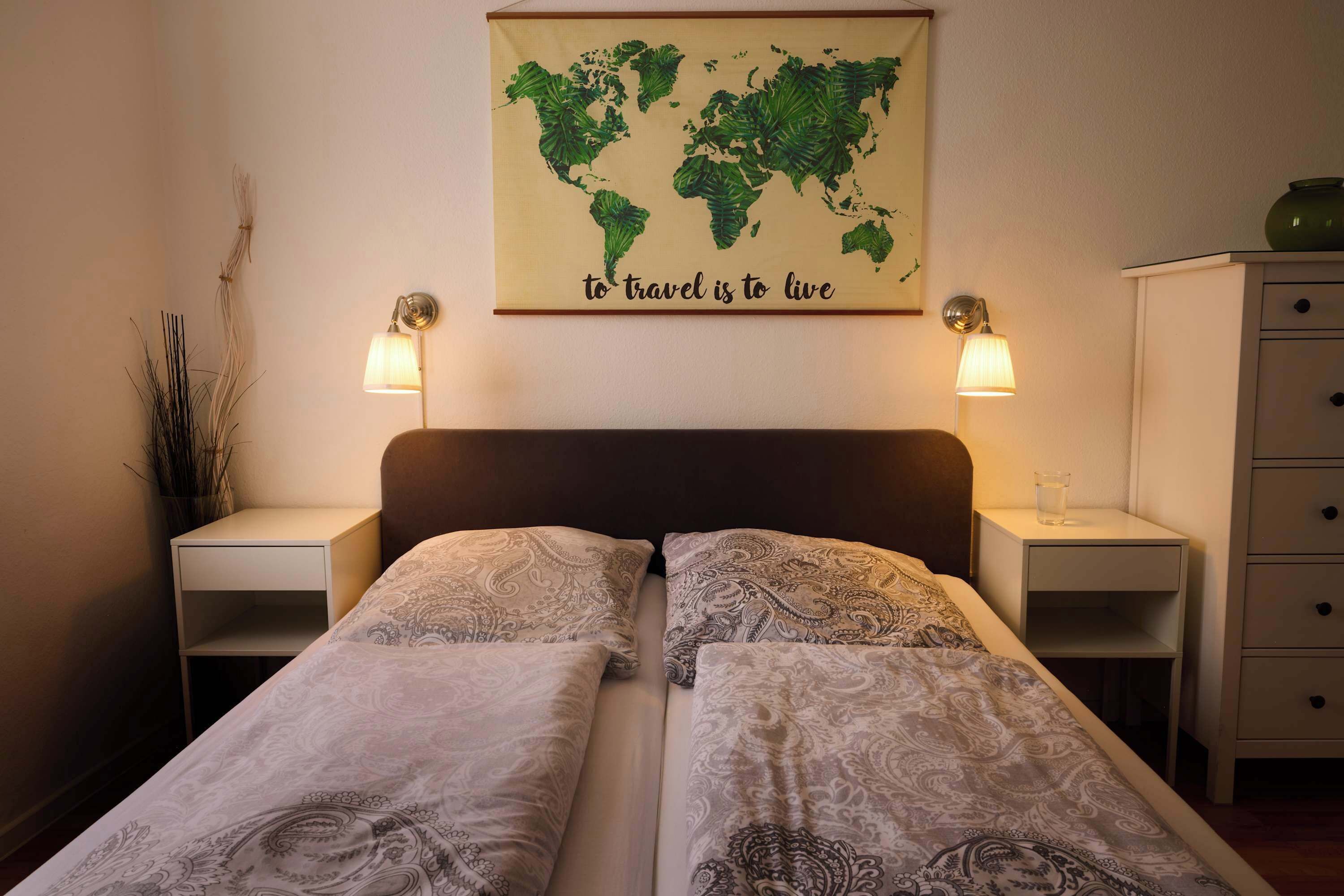 Una cama doble con ropa de cama estampada, dos lámparas de noche, un mapa del mundo con la frase 'to travel is to live' encima, y un mueble pequeño a la derecha.