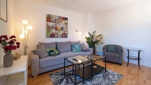 Ein Wohnzimmer mit grauem Sofa, einem Glastisch, einem kleinen Beistelltisch, einer Topfpflanze und einem Wandbild.