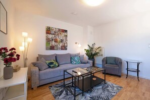 Ein Wohnzimmer mit grauem Sofa, einem Glastisch, einem kleinen Beistelltisch, einer Topfpflanze und einem Wandbild.