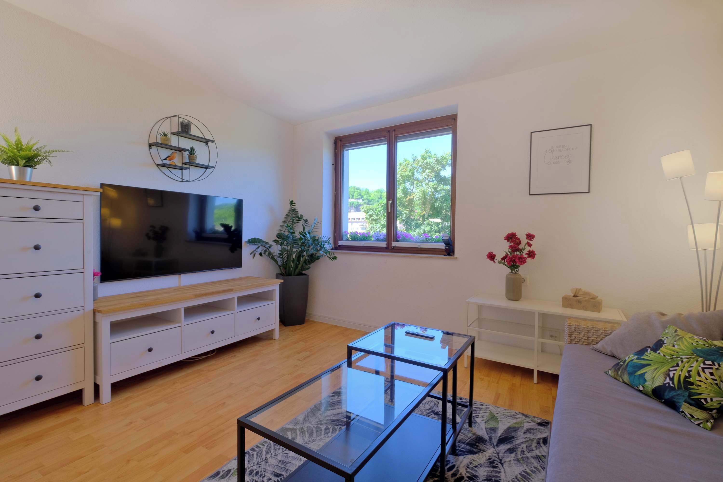 Een moderne woonkamer met een flatscreen televisie, een glazen salontafel, een bank met een decoratief plaid, een potplant en een raam met uitzicht op bomen.