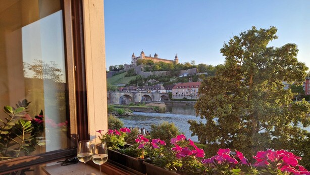 Blick aus dem Fenster auf einen Fluss, eine Burg auf einem Hügel und einen Balkon mit Blumen und Gläsern.