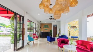 Un hall d’hôtel moderne avec un comptoir d’accueil, un espace salon avec des fauteuils rouges et bleus, et une grande fenêtre surmontée d’un auvent rouge.