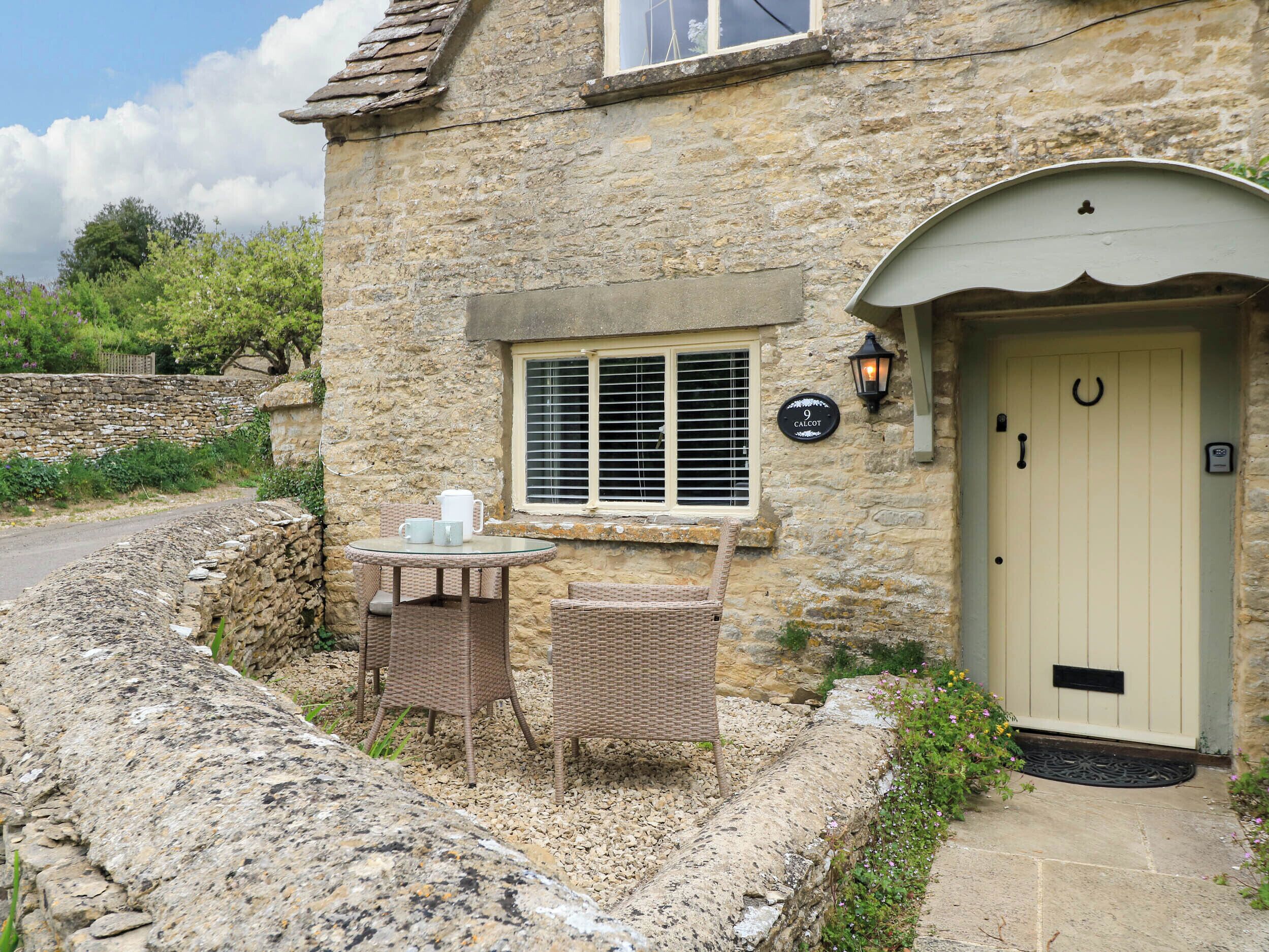 A stone building with a round table and wicker chairs outside, a door with a doormat, and a window with shutters.