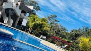 A swimming pool with clear blue water, surrounded by lush greenery and a modern building with balconies.