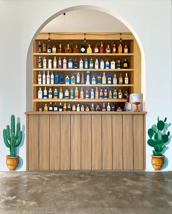 Un comptoir en bois, avec des étagères remplies de différentes bouteilles d’alcool, deux cactus en pot et une machine à café.