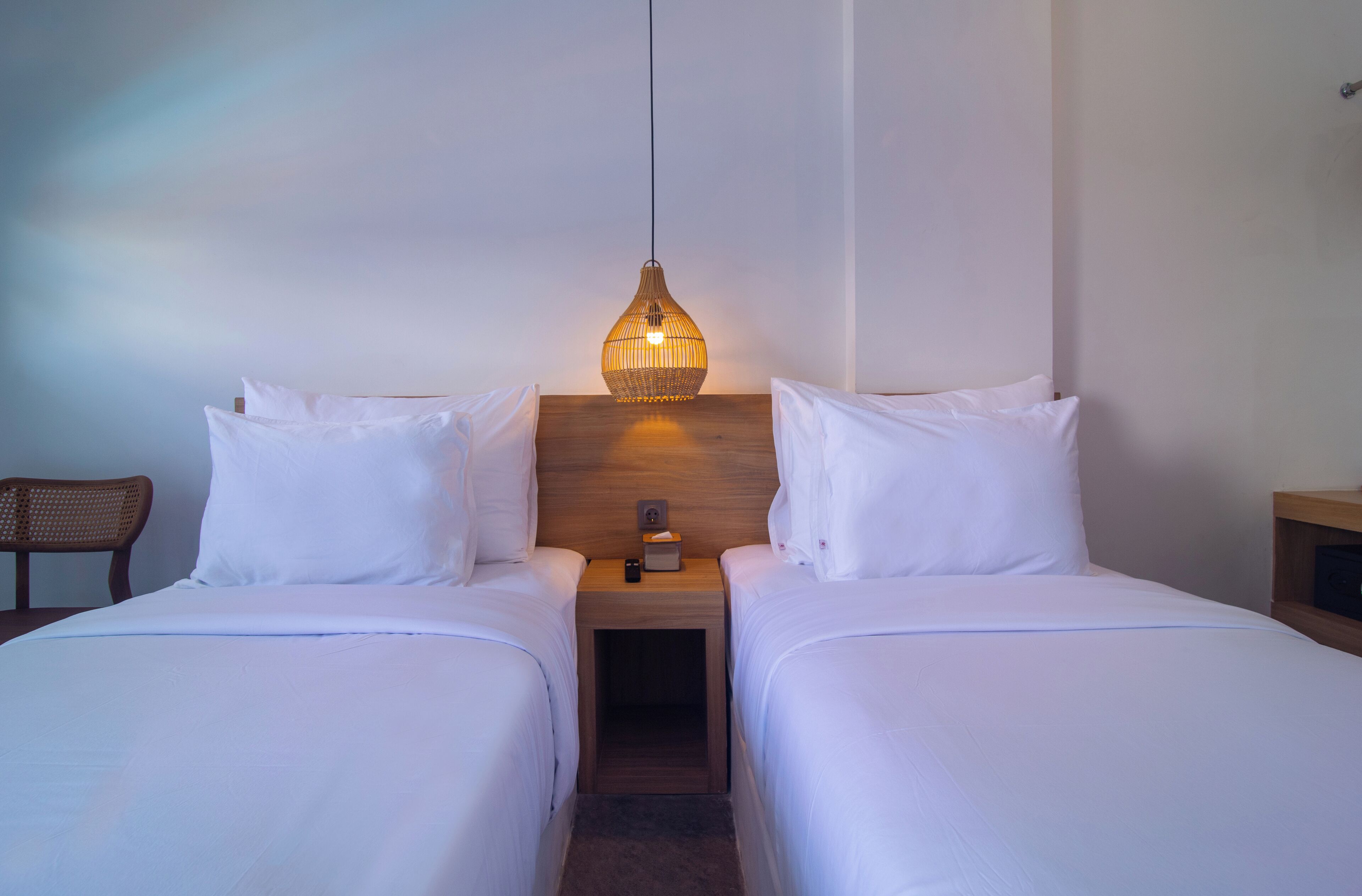 Habitación de hotel con dos camas individuales, cabecera de madera, mesita de noche con lámpara y una silla.