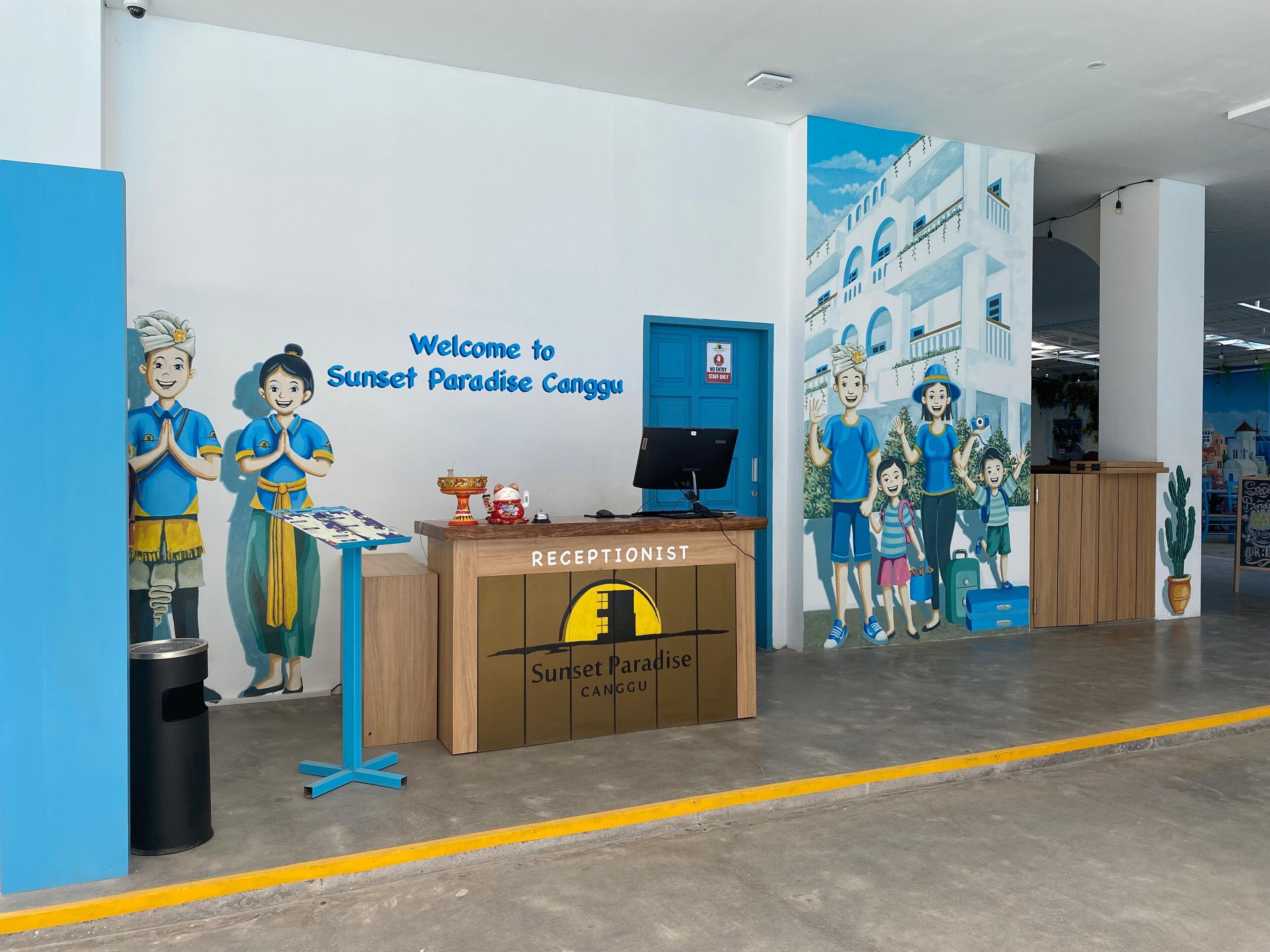A reception desk with a 'Welcome to Sunset Paradise Canggu' sign, a computer monitor, and a trash bin.