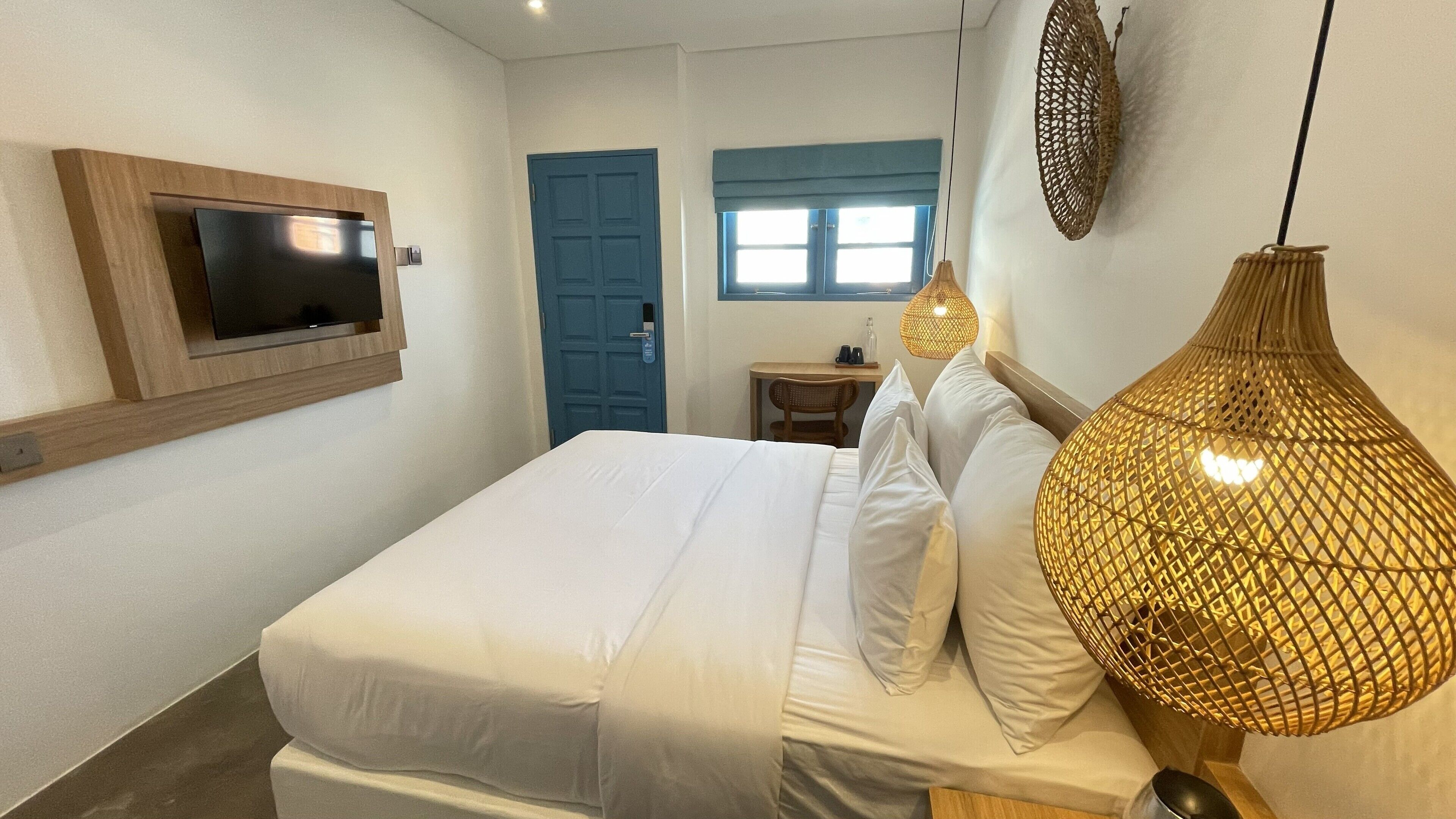 Ein Schlafzimmer mit einem Bett, einem an der Wand montierten Fernseher, einem Schreibtisch mit Stuhl und einem Fenster mit blauen Vorhängen.