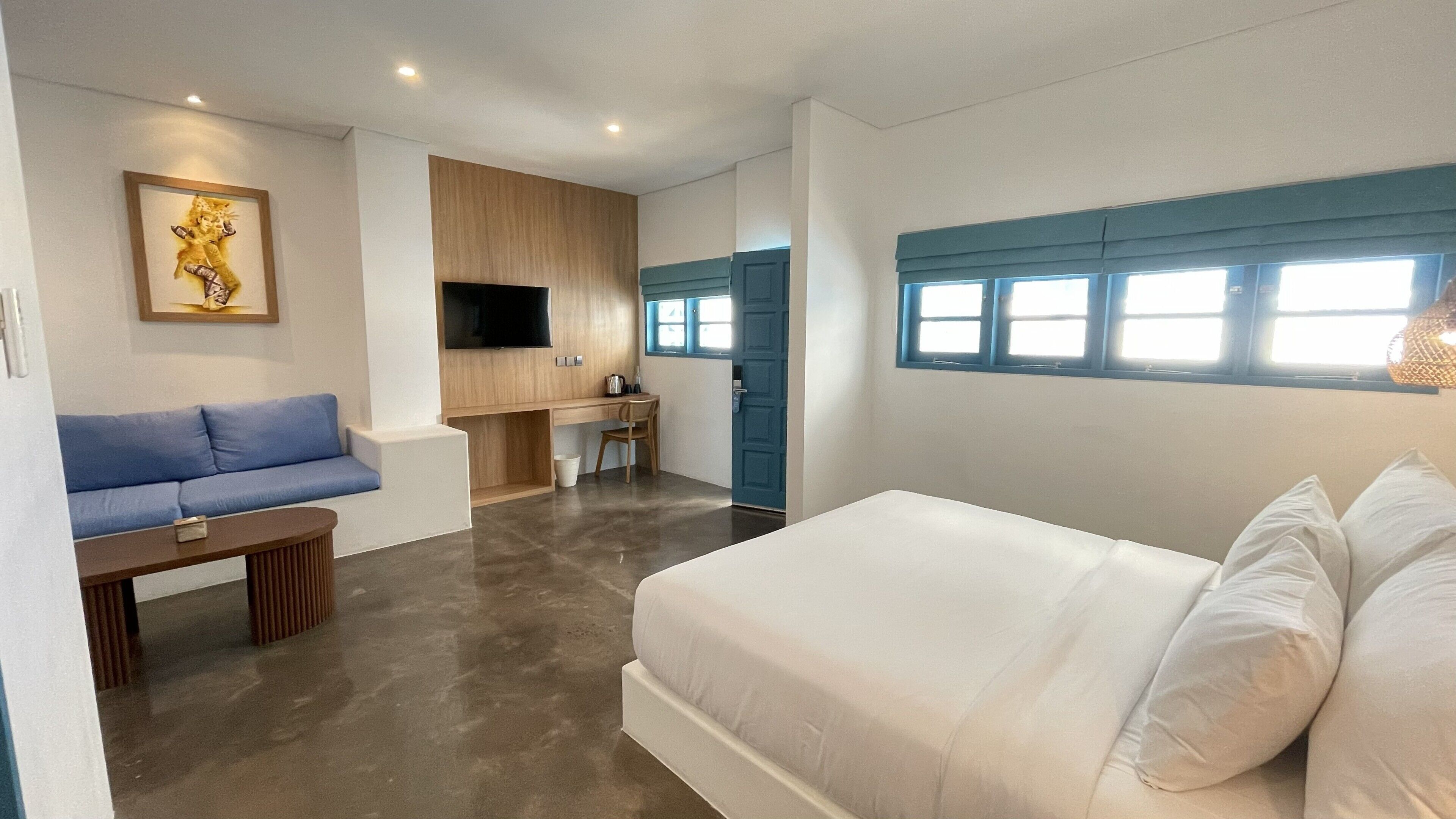A modern hotel room with a bed, a blue sofa, a desk with a chair, a television, and a window with blinds.