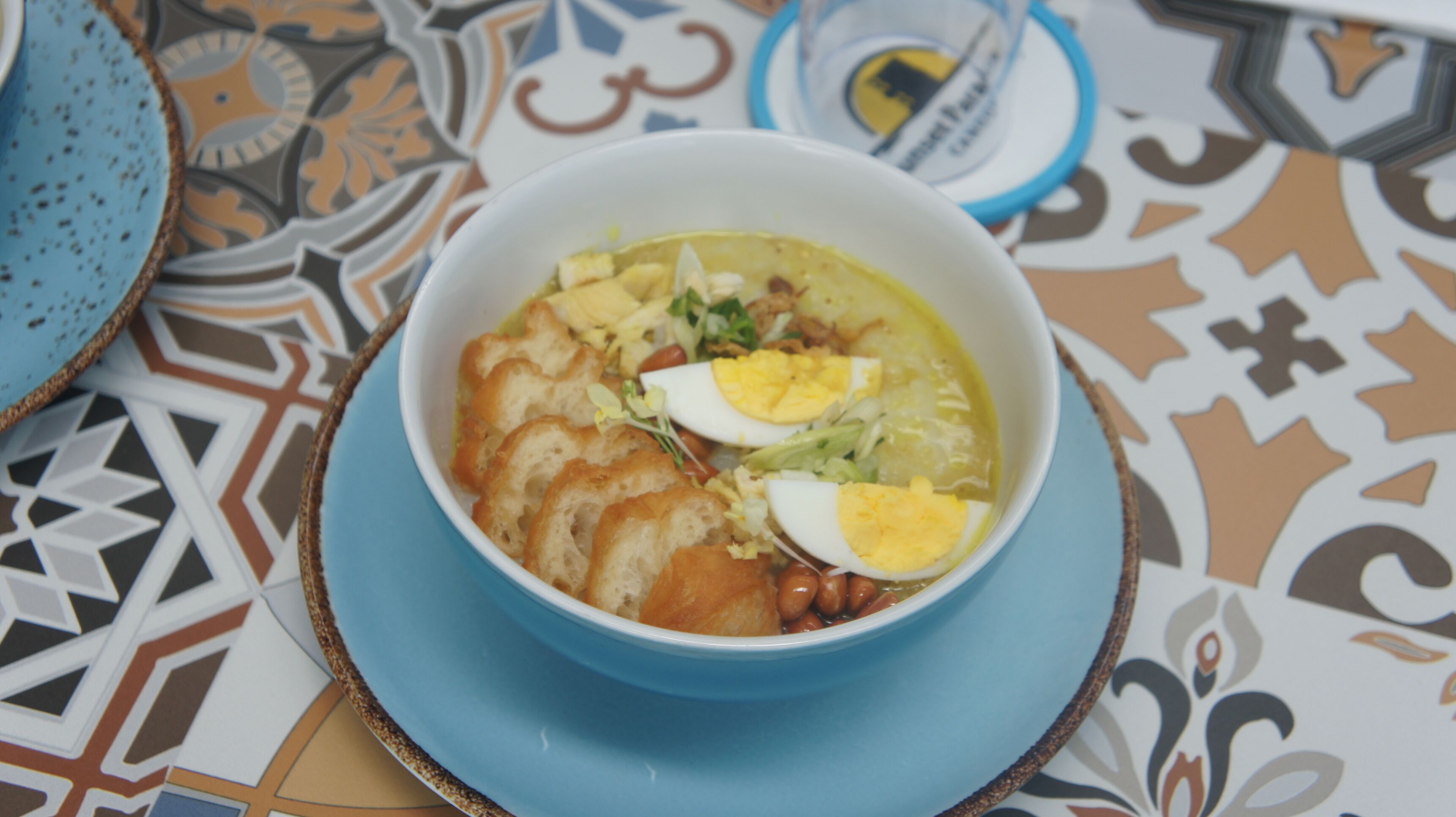 A bowl of soup with bread and eggs on a patterned tablecloth.