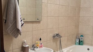A bathroom with a sink, mirror, and bathtub. There are toiletries on the sink and a trash bin next to the bathtub.