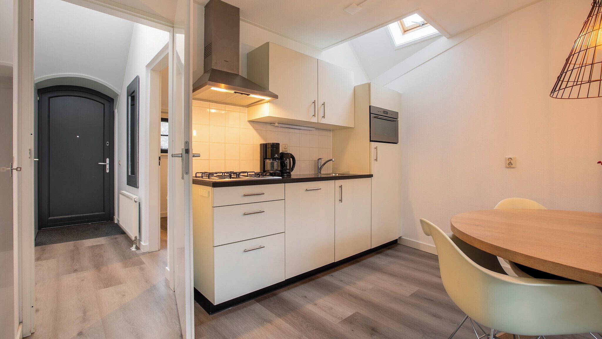 A compact kitchen with a gas stove, coffee maker, and a built-in oven. There is a dining area with a round table and two chairs. The kitchen has a skylight and a dark door.
