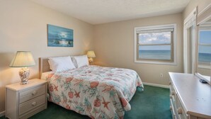 A bedroom with a bed, two bedside tables, a dresser, a painting, and a window with a view of the ocean. - GSH2 Ocean-Front Condo, Ocean Views, Comm.Pool (Kill Devil Hills)