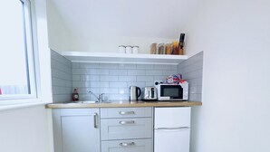 Une petite cuisine avec des meubles blancs, un évier, un grille-pain, un four à micro-ondes et un petit réfrigérateur. Il y a une fenêtre sur le côté gauche et une poubelle sur le sol.