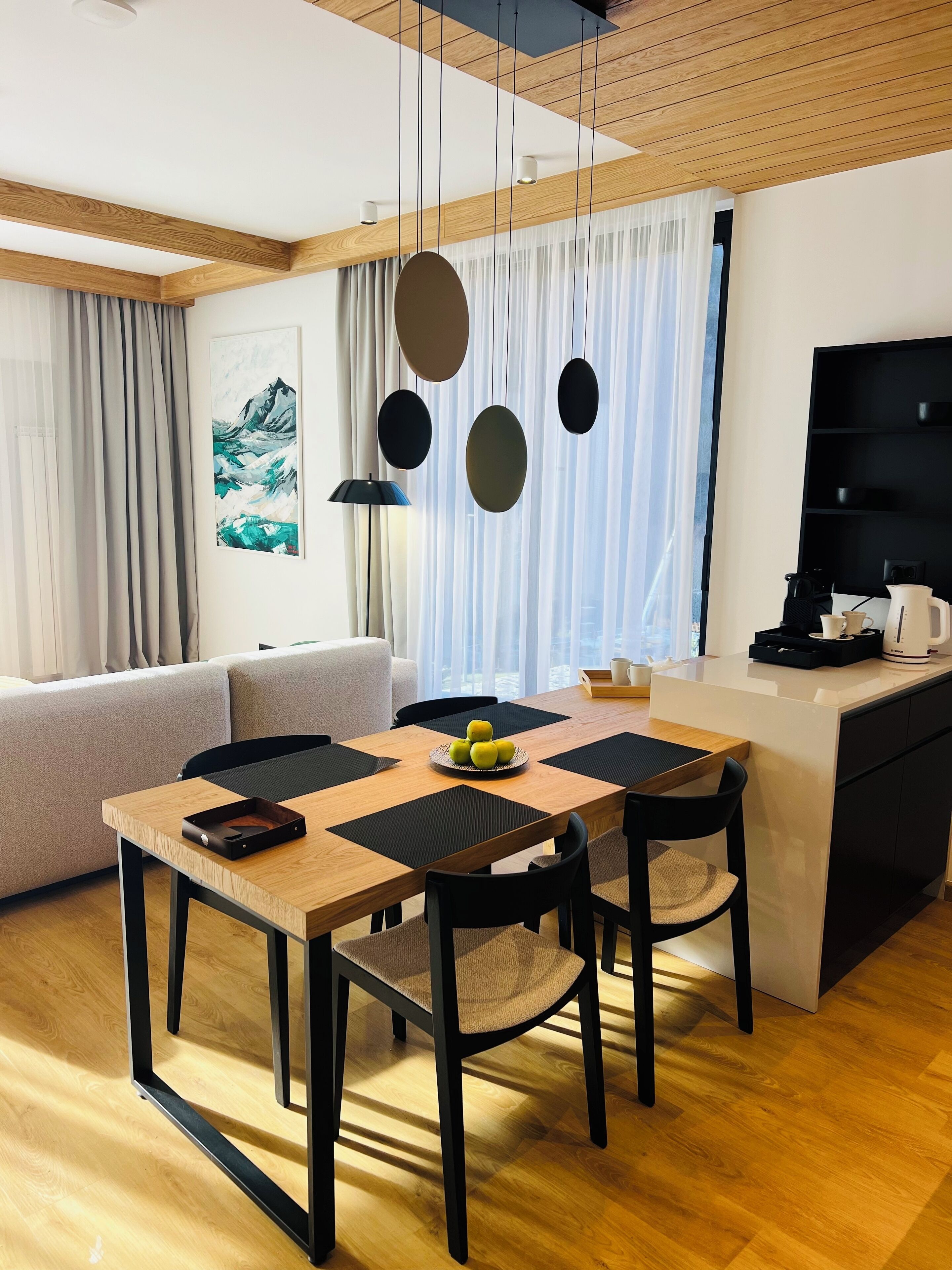 A modern living room with a wooden dining table, black chairs, and a sofa. There is a kitchen area with a black countertop and a coffee machine.