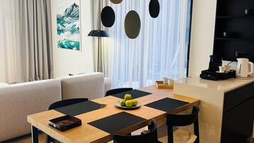 A modern living room with a wooden dining table, black chairs, and a sofa. There is a kitchen area with a black countertop and a coffee machine.