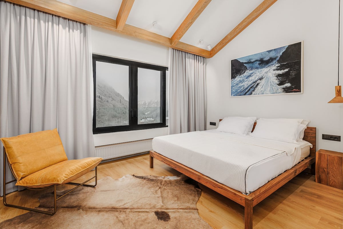 Un dormitorio con cama de madera, una silla amarilla, un ventanal grande y vistas a la montaña.