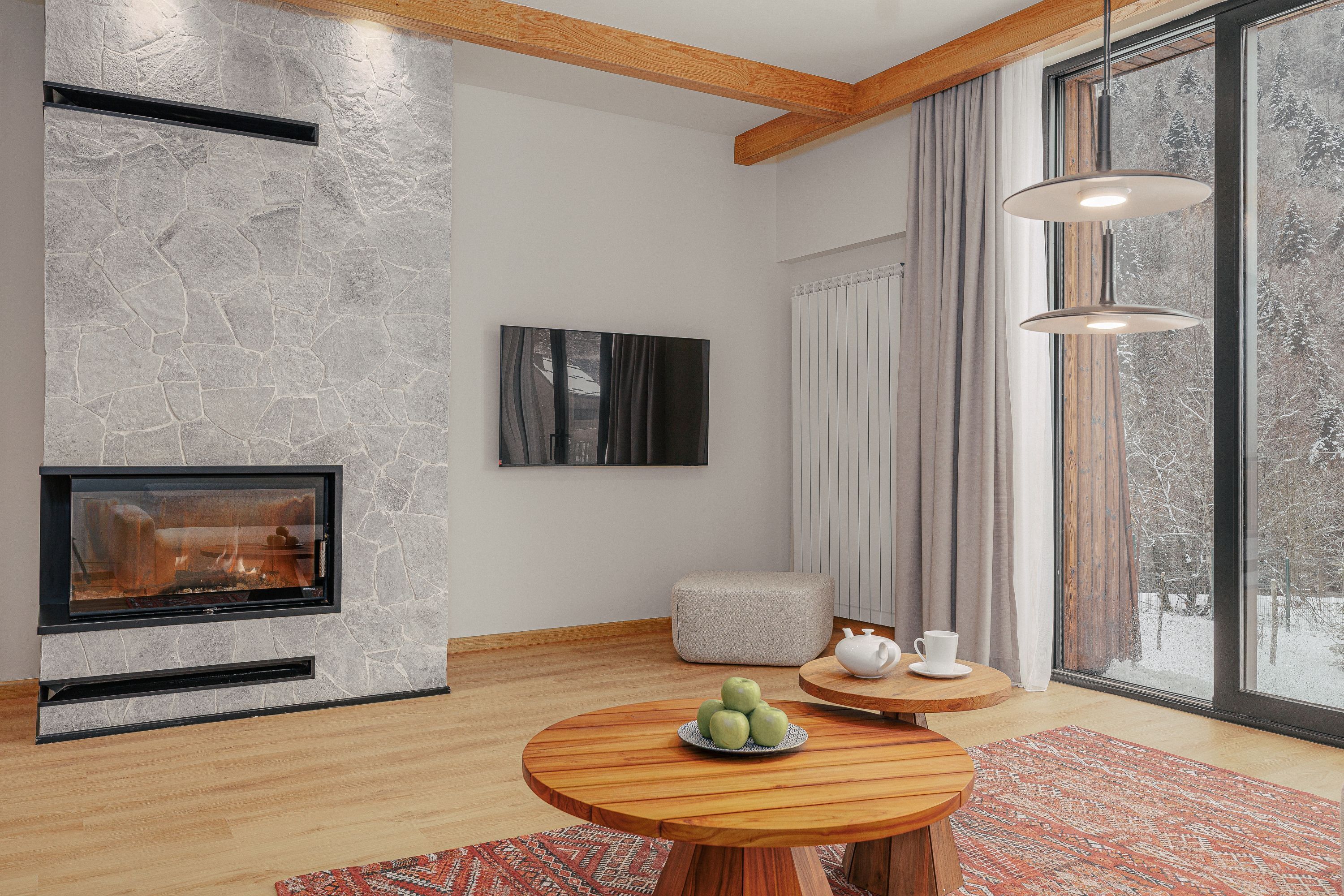A modern living room with a stone fireplace, a round wooden table with a fruit bowl, a white pouf, and a view of a snowy landscape through large windows.