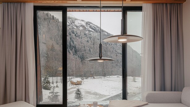 Une pièce chaleureuse avec une grande fenêtre donnant sur un paysage montagneux enneigé, un canapé, une petite table ronde avec une théière et une tasse, et un luminaire suspendu.