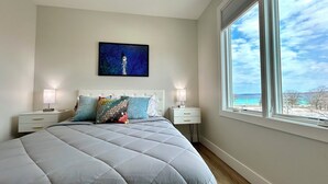 A bedroom with a large bed, two bedside tables, a nightstand, a window with a view of the sea, and a painting on the wall. - The LUX~ 1 Block to the Beach!! (Traverse City)