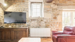 Une pièce aux murs de pierre, avec un téléviseur fixé au mur, une table en bois, un canapé en cuir rouge et une fenêtre donnant sur un paysage agréable.