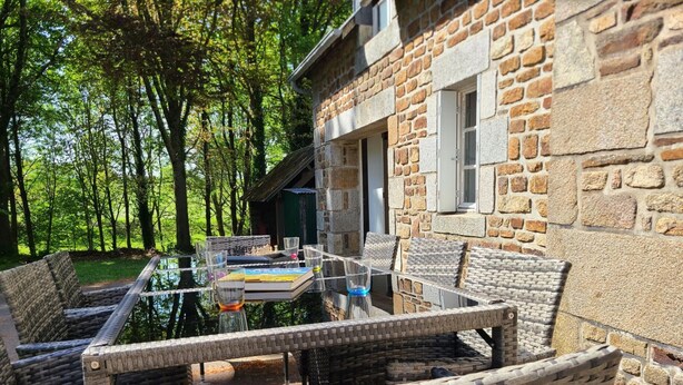 Un espace de restauration en plein air, avec des chaises en osier et une table en verre, situé devant un bâtiment en pierre et offrant une vue sur les arbres.