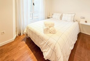 A neatly made bed with a white quilt and rolled towels, a wooden floor, a nightstand with a lamp, and a window with curtains. - XILGAR Apartment Av Leon on the Beach (Sanxenxo)