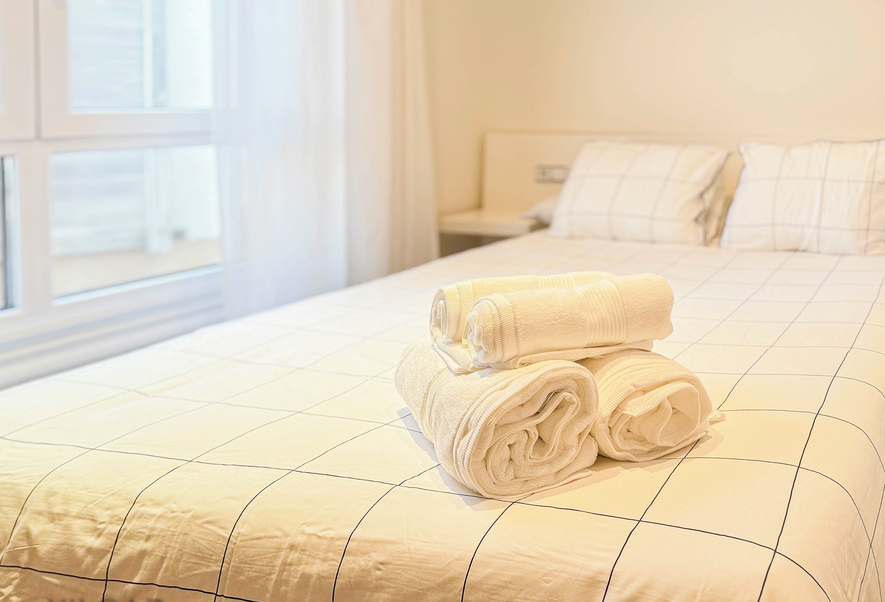 A neatly made bed with rolled white towels, a quilted bedspread, and a window with curtains.